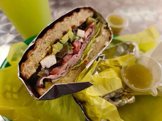 Wrapped sandwich with lettuce, tomato, and cheese beside a small container of sauce on yellow paper