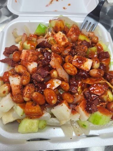 Takeout container of diced fruit or dessert topped with chocolate, peanuts, and whipped cream, with a fork