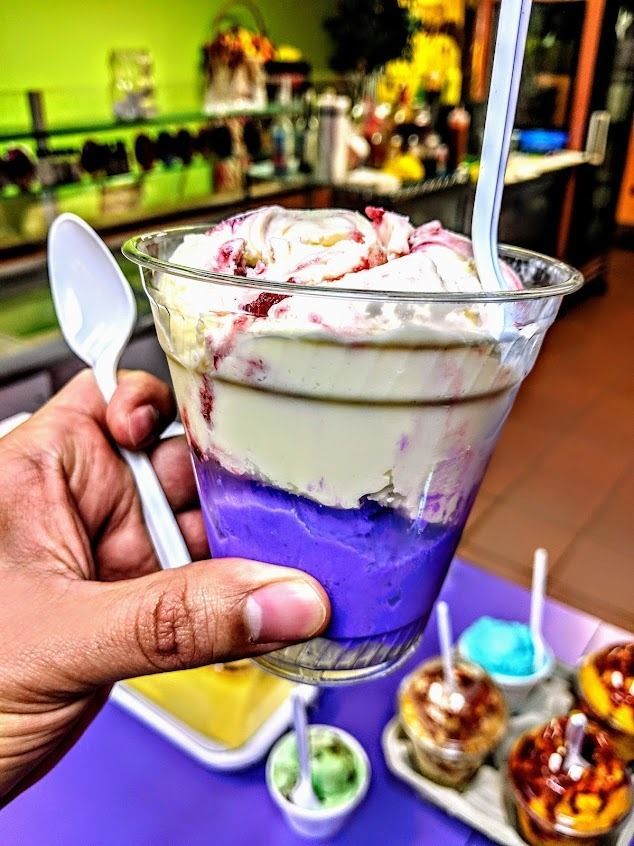 Hand holding a layered purple and vanilla dessert in a clear cup with a spoon at a café counter