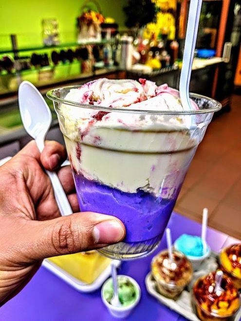 Hand holding a purple-and-cream layered ice cream sundae in a clear cup with a spoon and wafer stick.