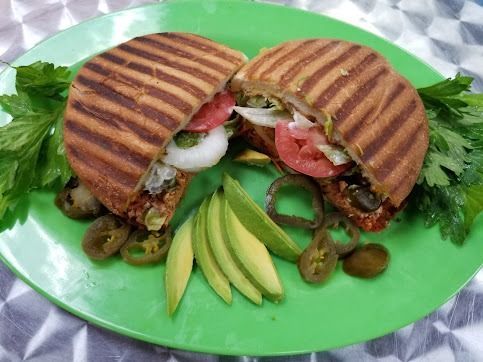 Grilled sandwich halves on a green plate with lettuce, tomato, onion, olives, and avocado slices