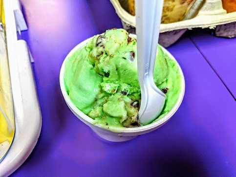 Green ice cream in a cup with a spoon on a purple table, with other dessert cups nearby