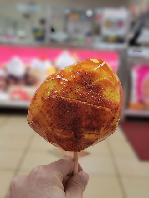 Hand holding a glossy orange-red candied fruit on a stick in a blurred store aisle