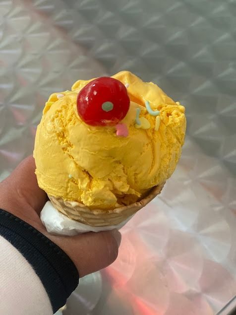 Yellow ice cream cone topped with a red cherry, held in a hand against a metal table background
