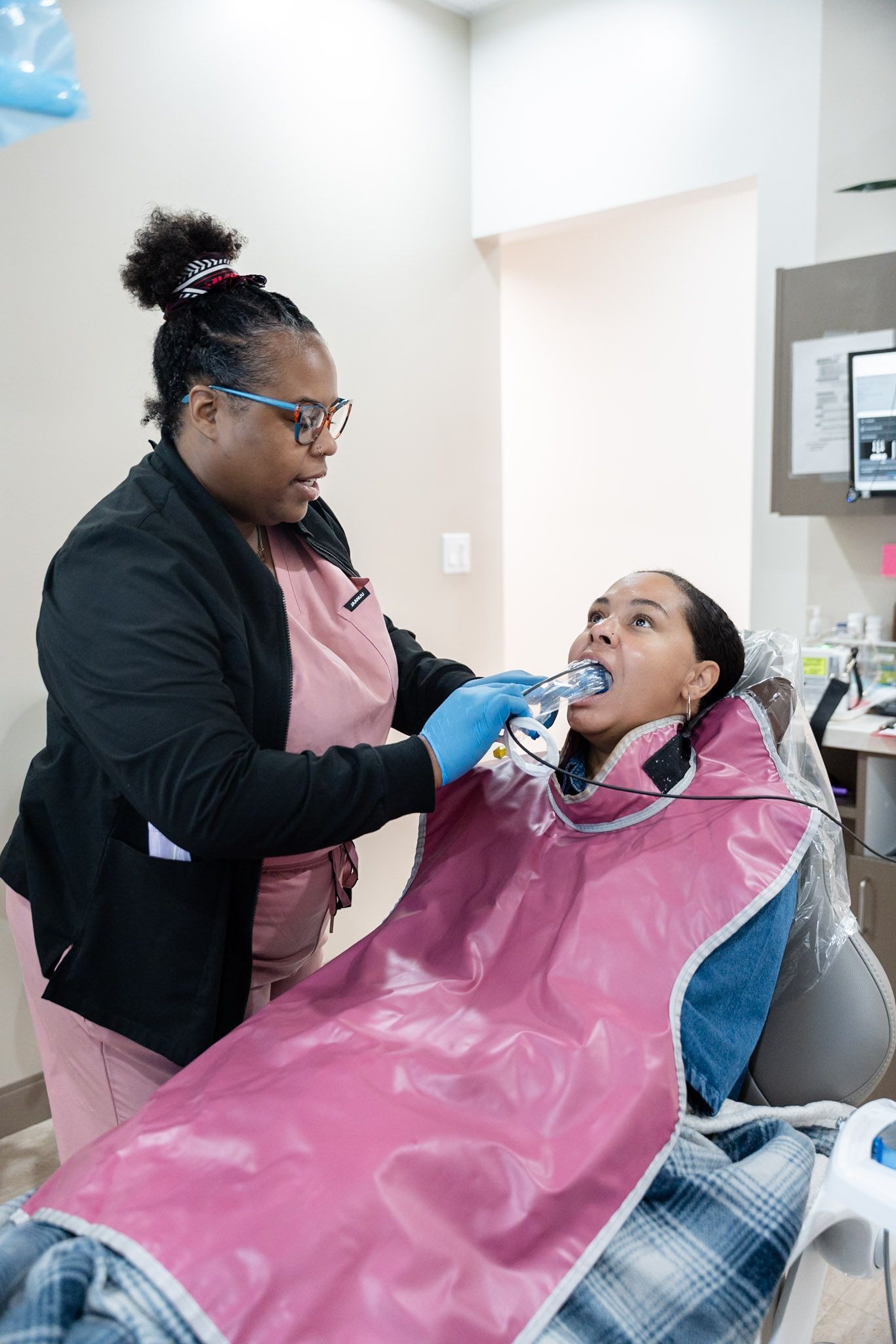Dental hygienist treating a patient in a chair, wearing pink bib and blue gloves.