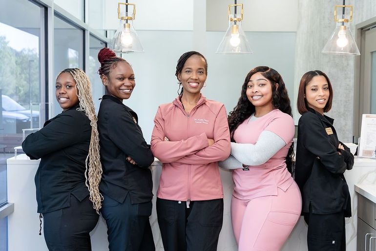 Five people posing together in a bright office, smiling with arms crossed, wearing black, pink, and white outfits
