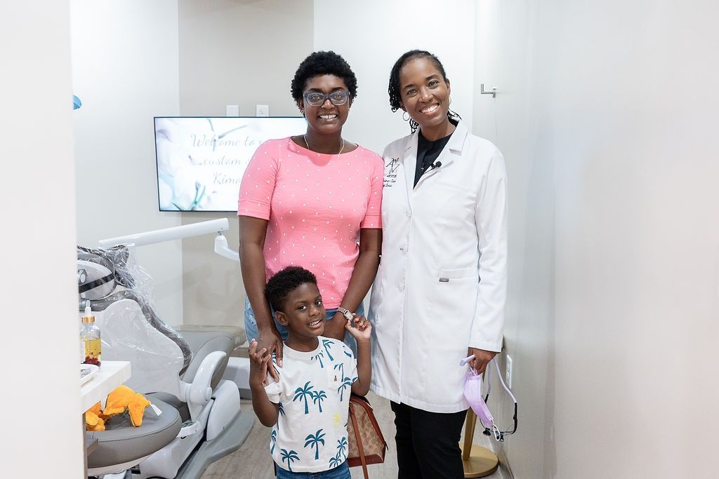 Two smiling adults and a child standing in a clinic room beside a medical chair
