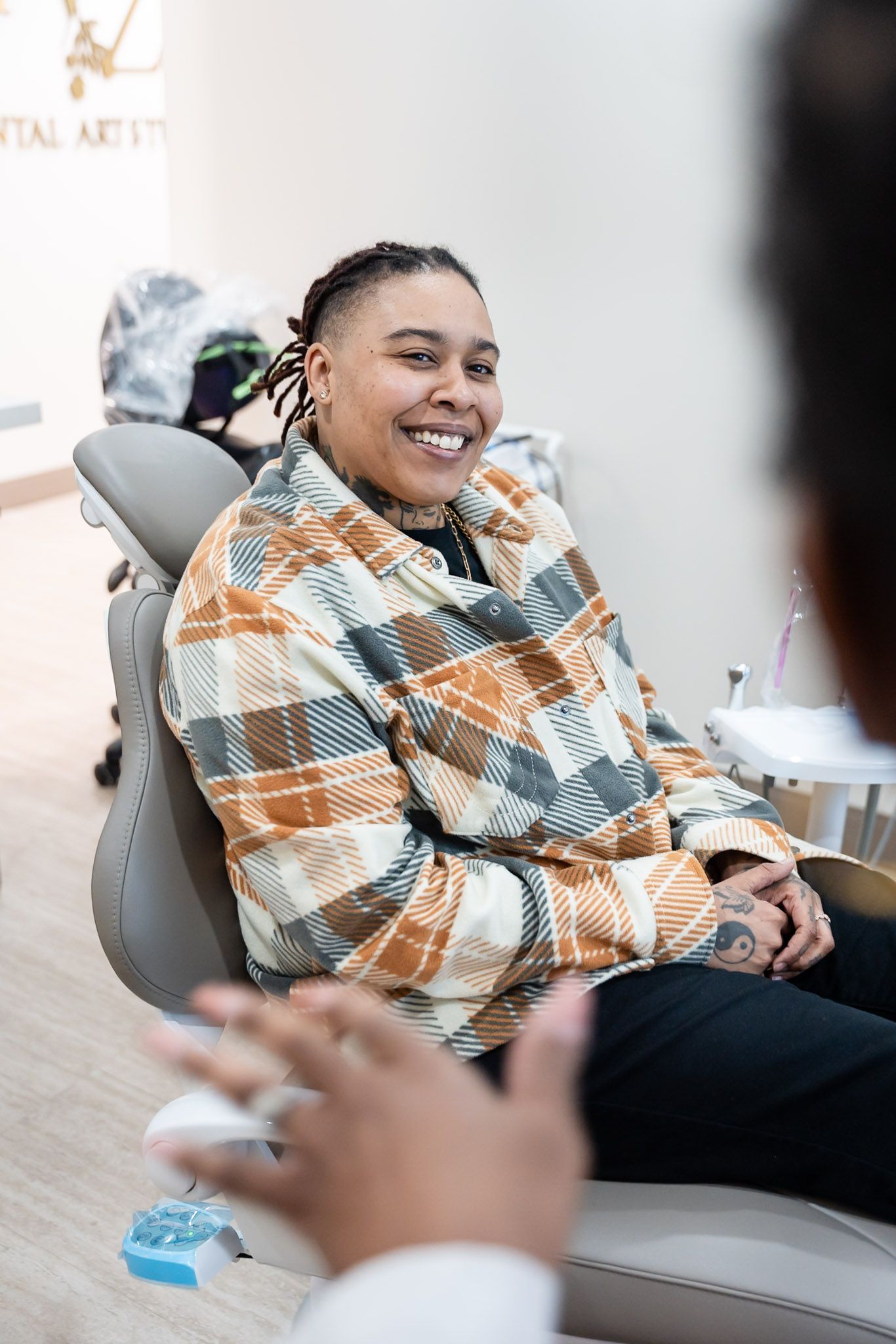 Smiling person sitting in a dental chair, wearing a plaid blanket in a bright clinic.