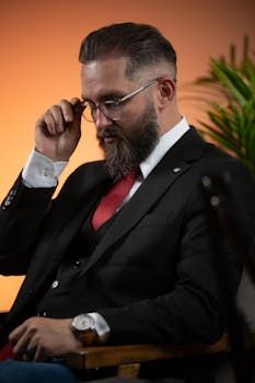 Un homme en costume et cravate est assis sur une chaise et ajuste ses lunettes.