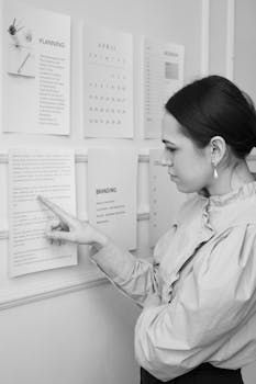 Une personne en chemise à col désigne un document épinglé au mur, entouré d'autres documents de planification.