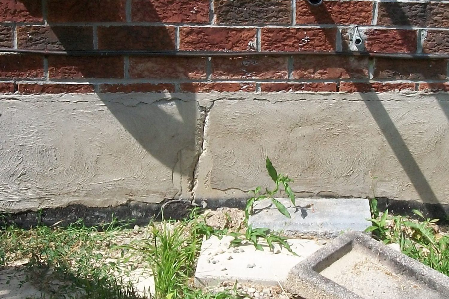 Brick wall with a cracked concrete base and weeds growing along the ground.