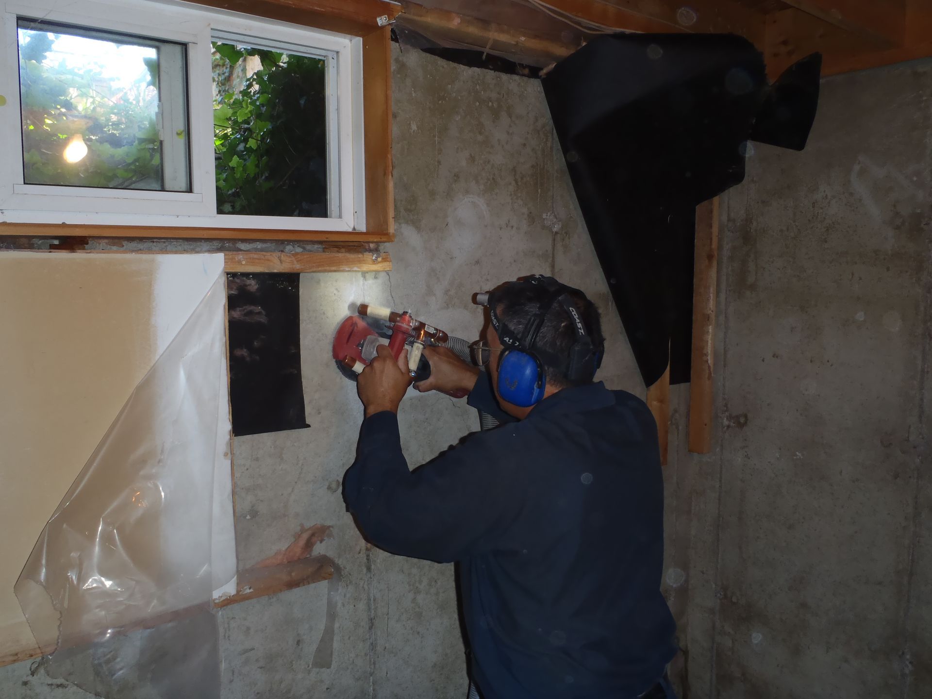 Person using a power drill on a wall in a partially finished room, wearing earmuffs and a respirator.