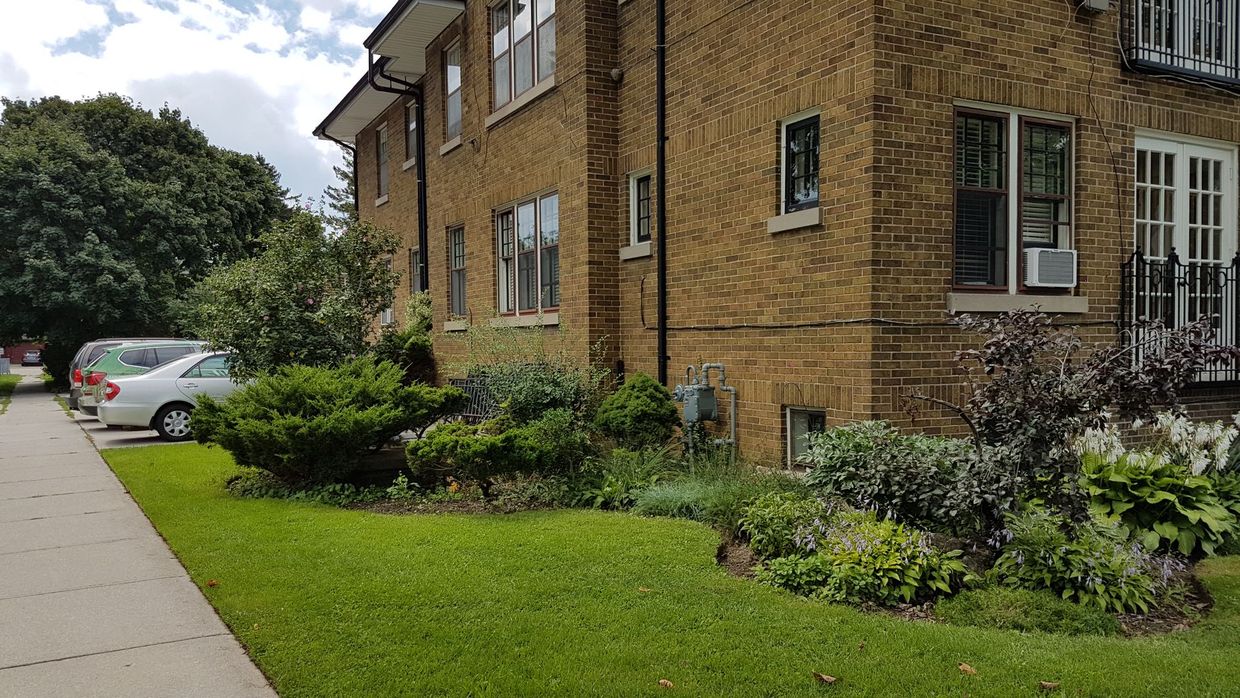 Brick apartment building beside a green lawn, shrubs, and a sidewalk with parked cars nearby