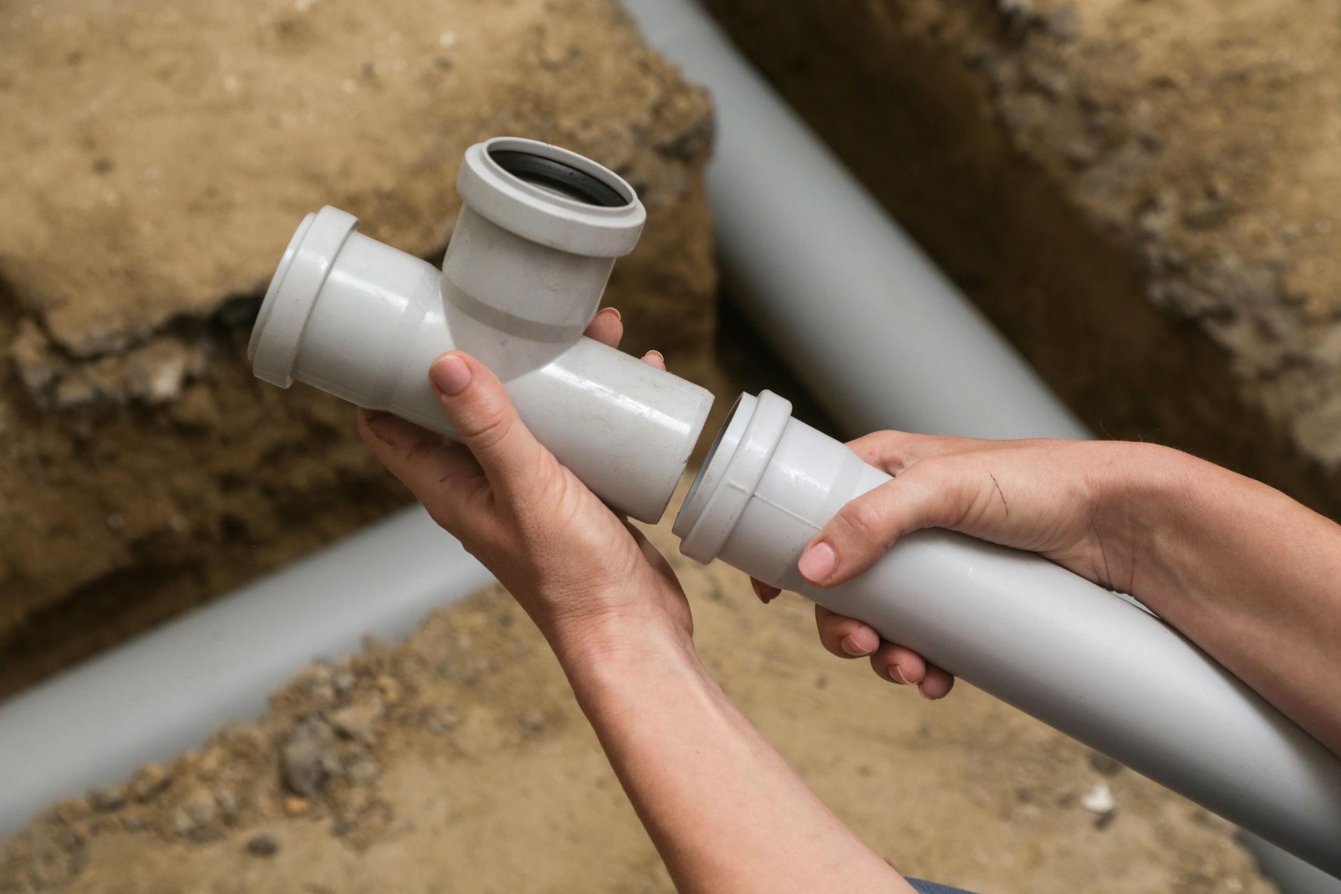 Hands connect white PVC pipes over a dirt trench during plumbing installation.