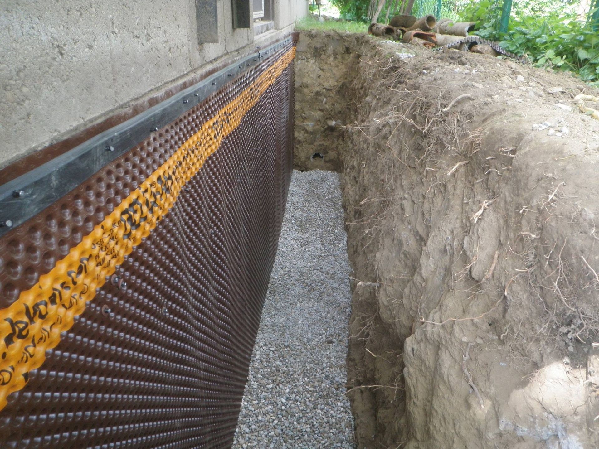 Foundation wall with black drainage mat and orange membrane beside a deep trench outdoors