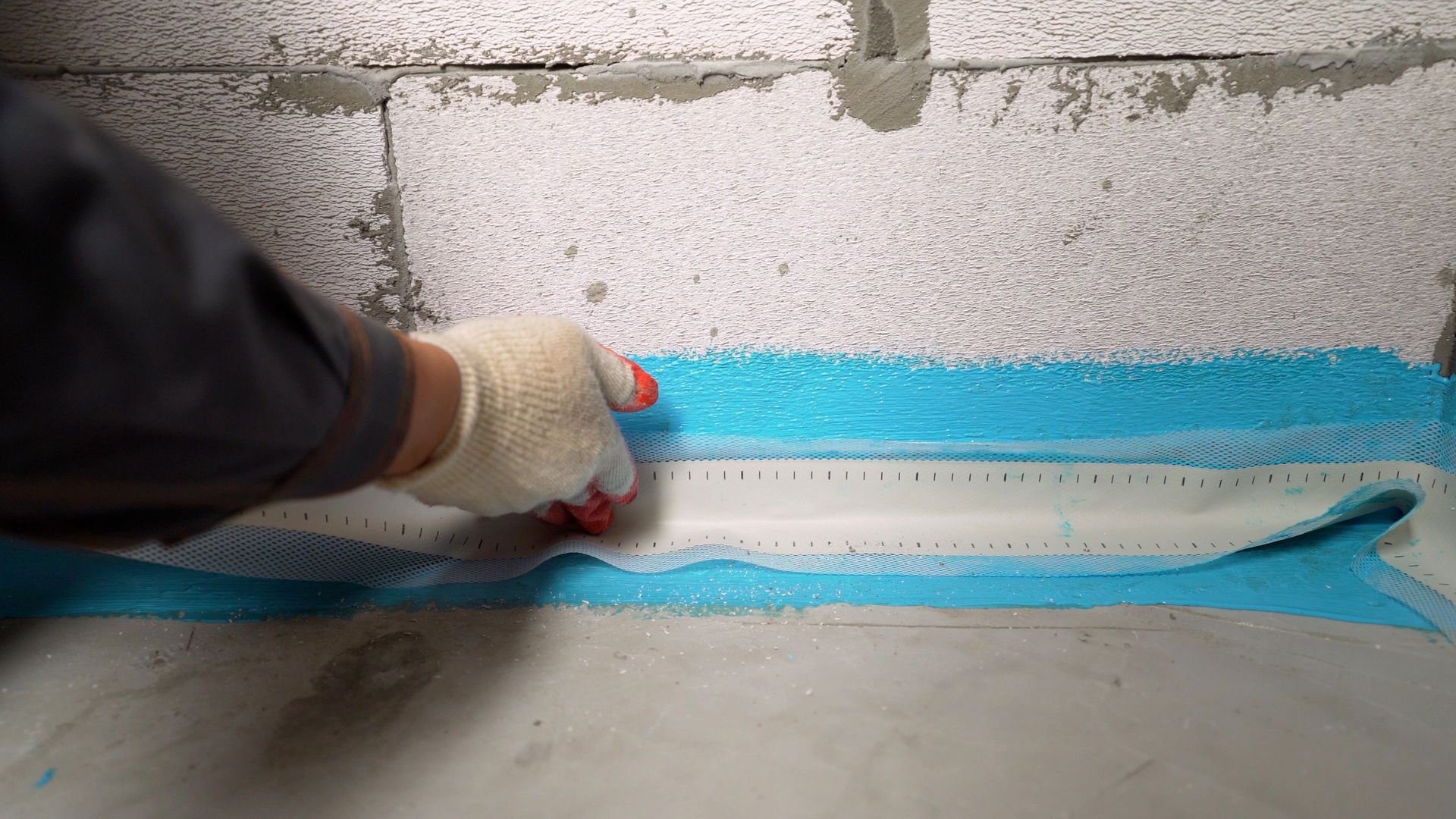 A gloved hand applies blue waterproofing membrane to a concrete surface, against a cinder block wall.