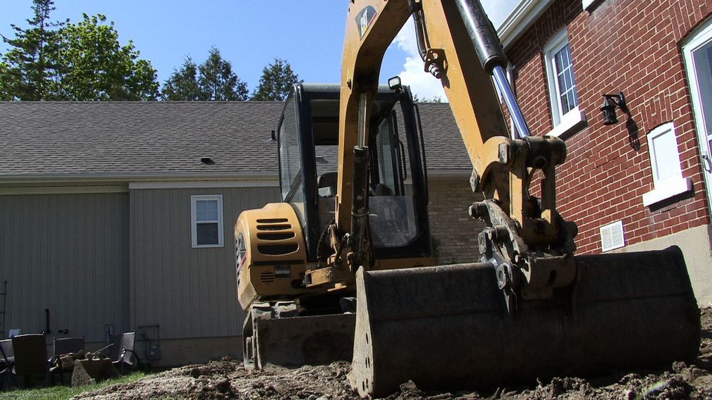 Yellow excavator beside a red brick house and shed in a backyard under a clear blue sky