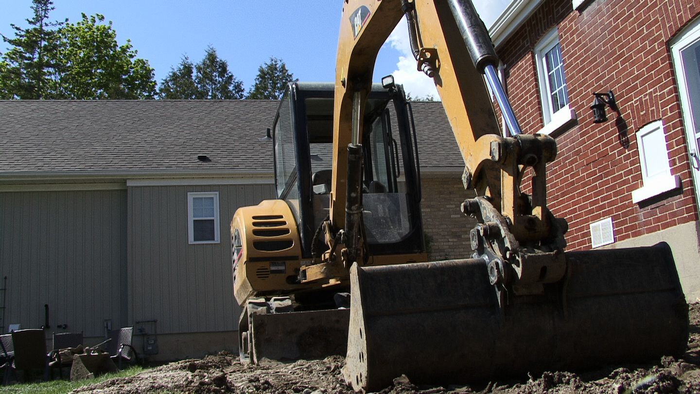 Yellow excavator beside a red brick house and shed in a backyard under a clear blue sky