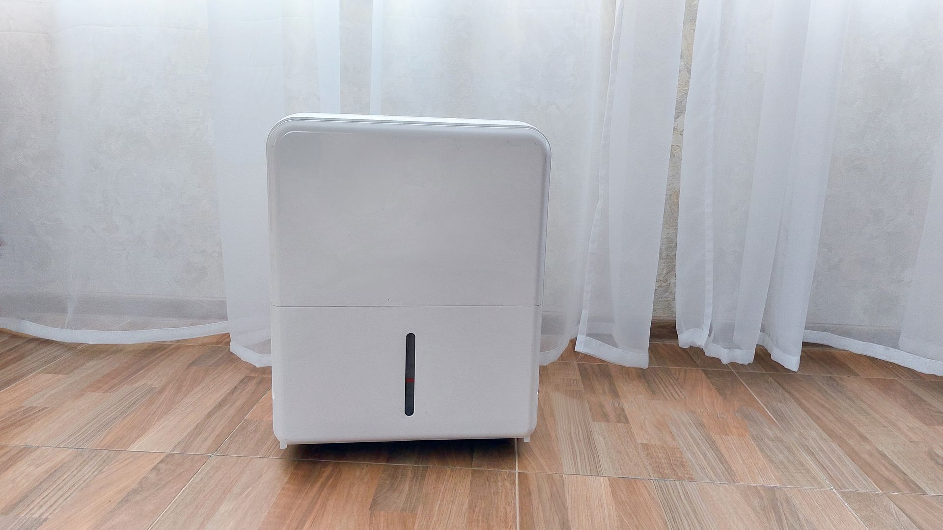 White portable appliance centered on a wood floor in front of sheer curtains.