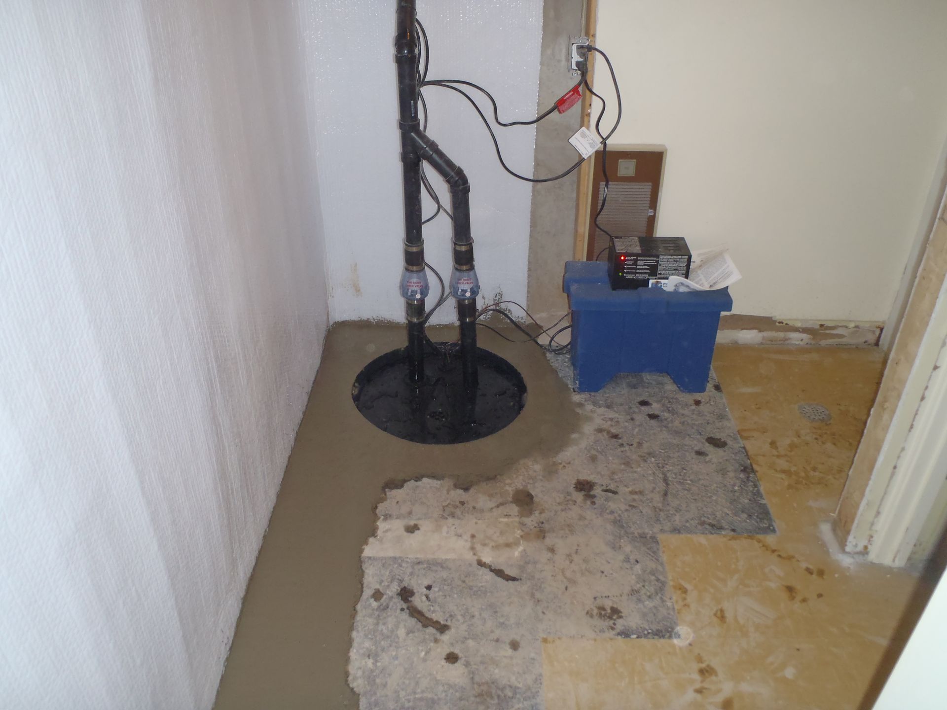 Sump pump installation in a basement corner, with a black pipe, blue box, and wet concrete.