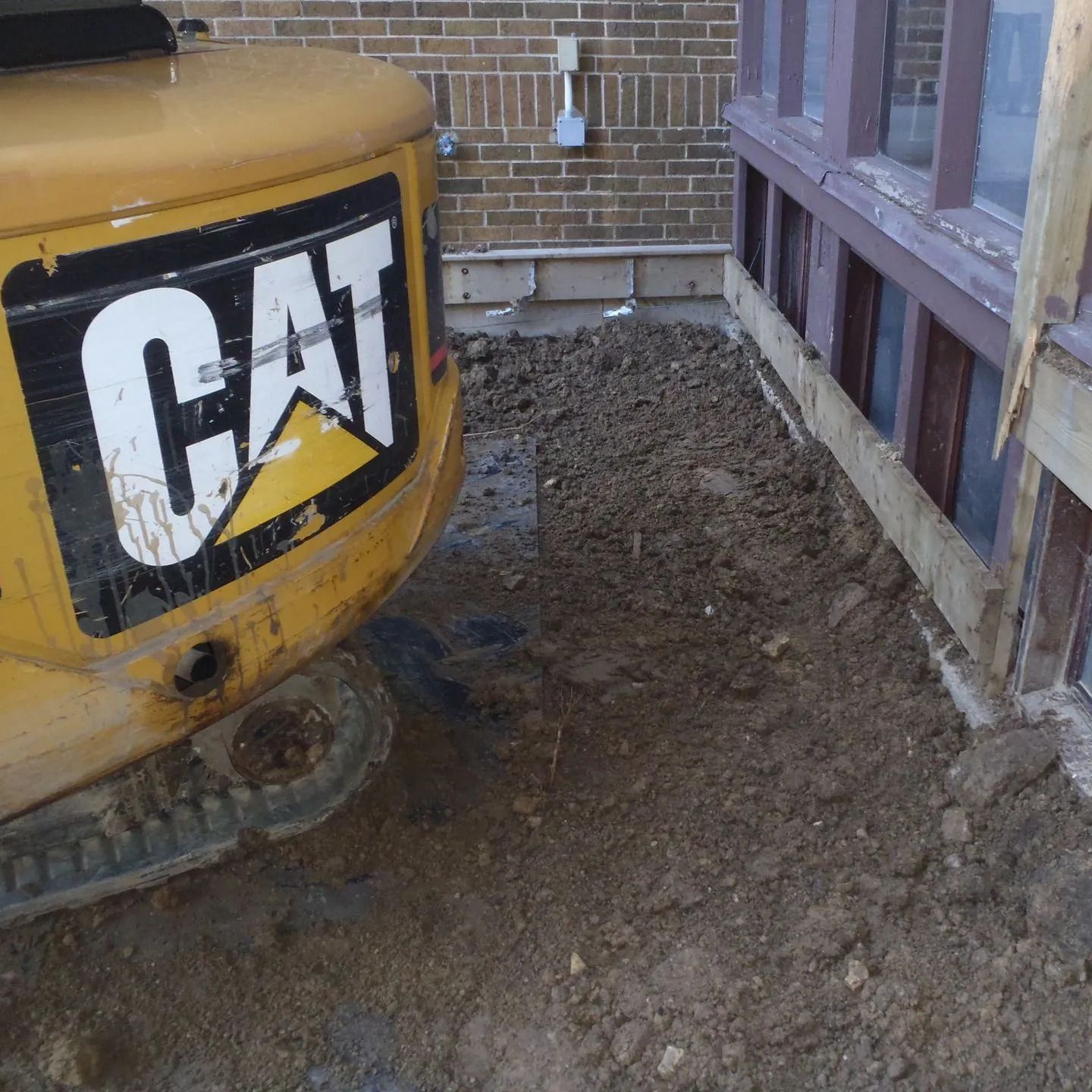 Yellow CAT excavator beside a brick building on a dirt construction site.