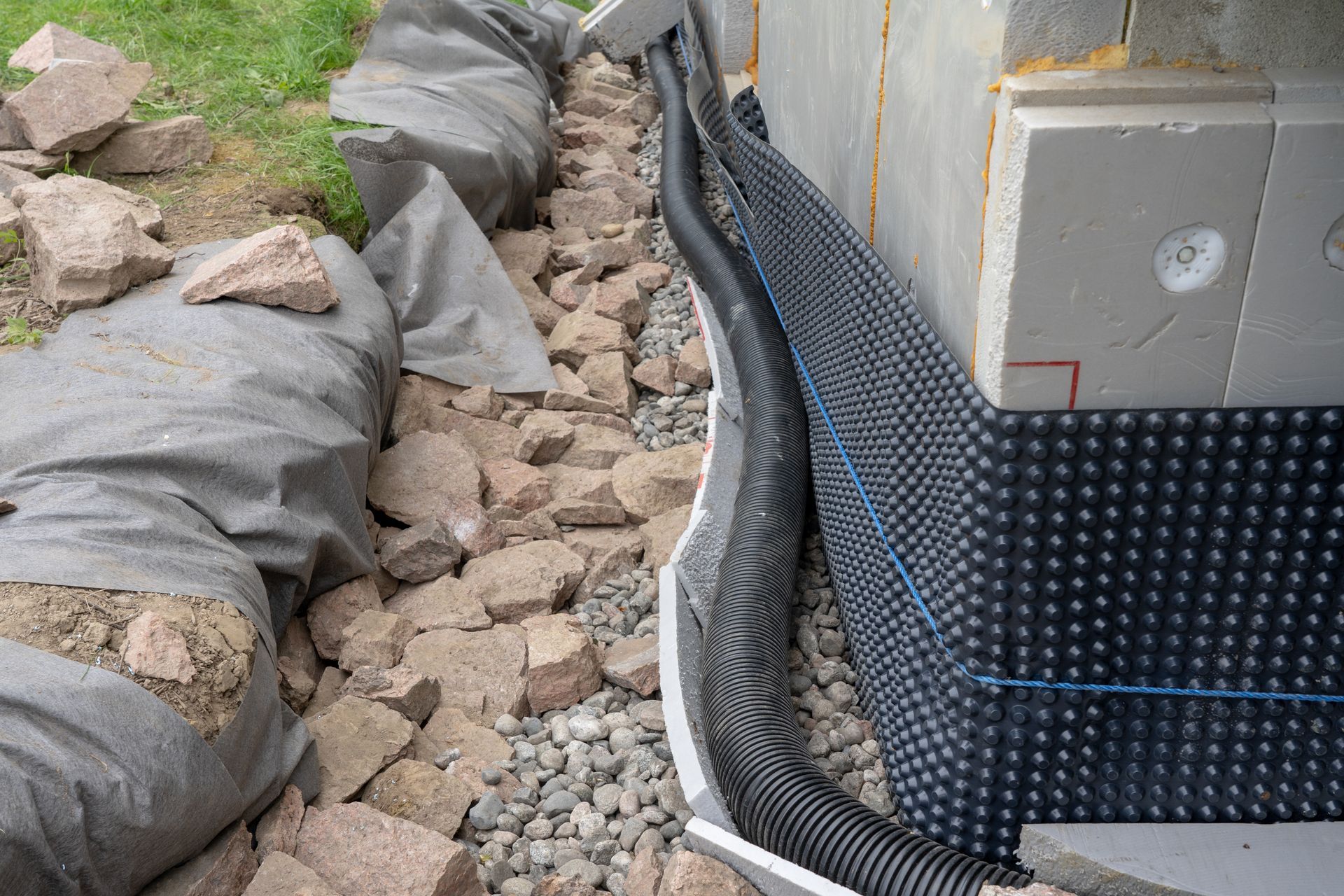 Foundation wall with black drain mat, corrugated pipe, and gravel beside landscape fabric under construction