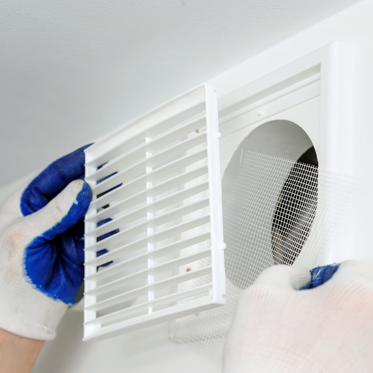 Hands in gloves removing a white air vent cover from a wall.