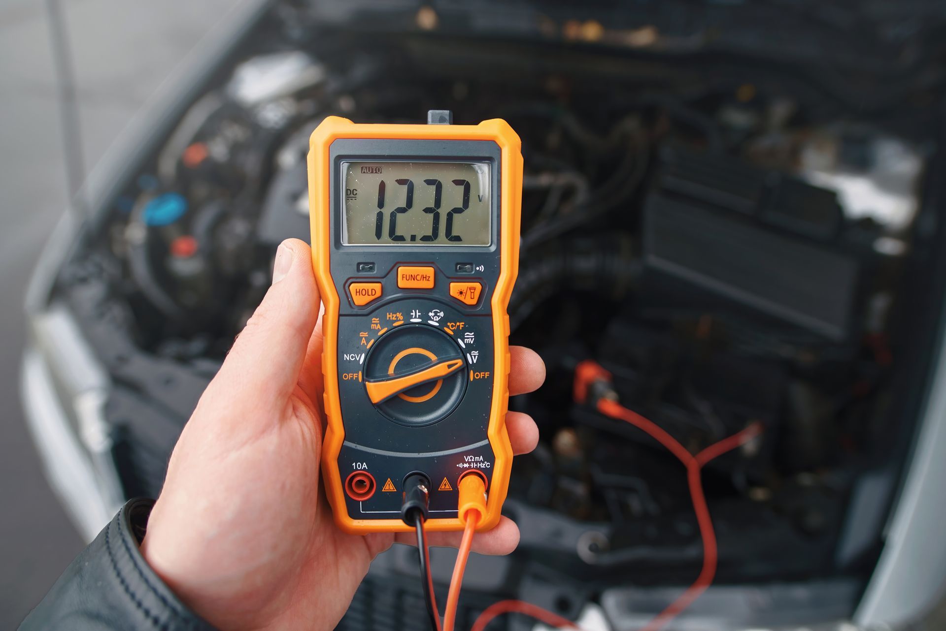 Hand holding multimeter showing 12.32 volts, testing a car battery in an open engine bay.