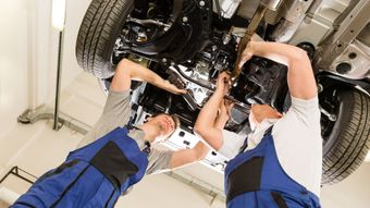 Mechanics working on the underside of a car in a garage, wearing blue overalls.