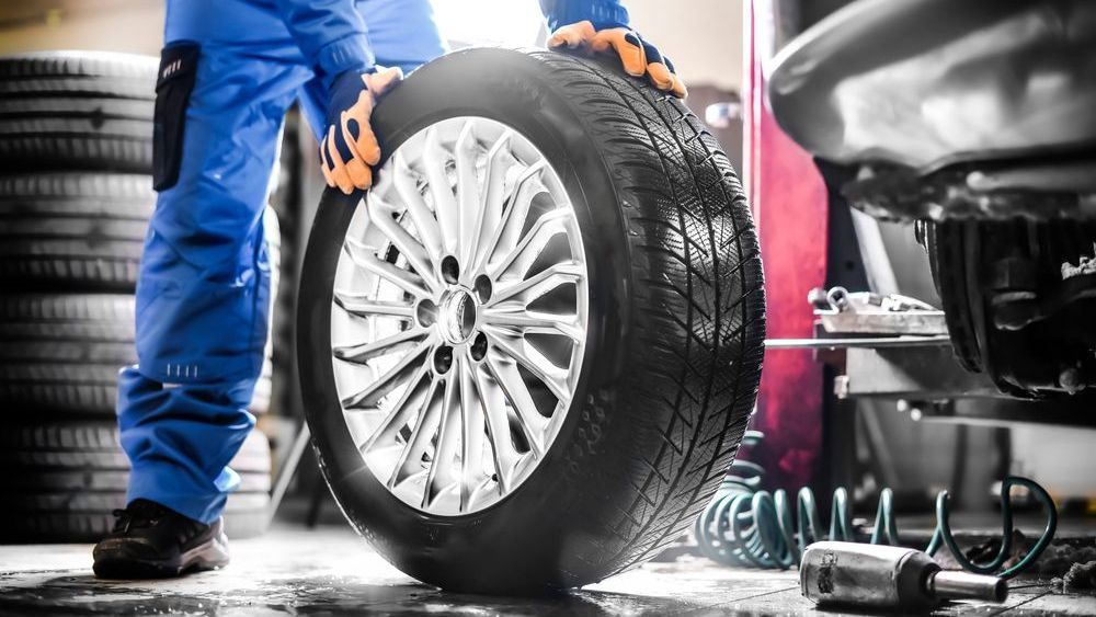 Mechanic holding a car tire in a garage, wearing blue coveralls and gloves.