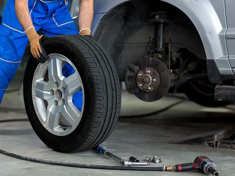 Mechanic in blue jumpsuit changing a car tire; tire off the vehicle, tools visible.