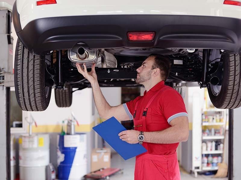 Mechanic inspecting car exhaust while holding a clipboard in a repair shop.