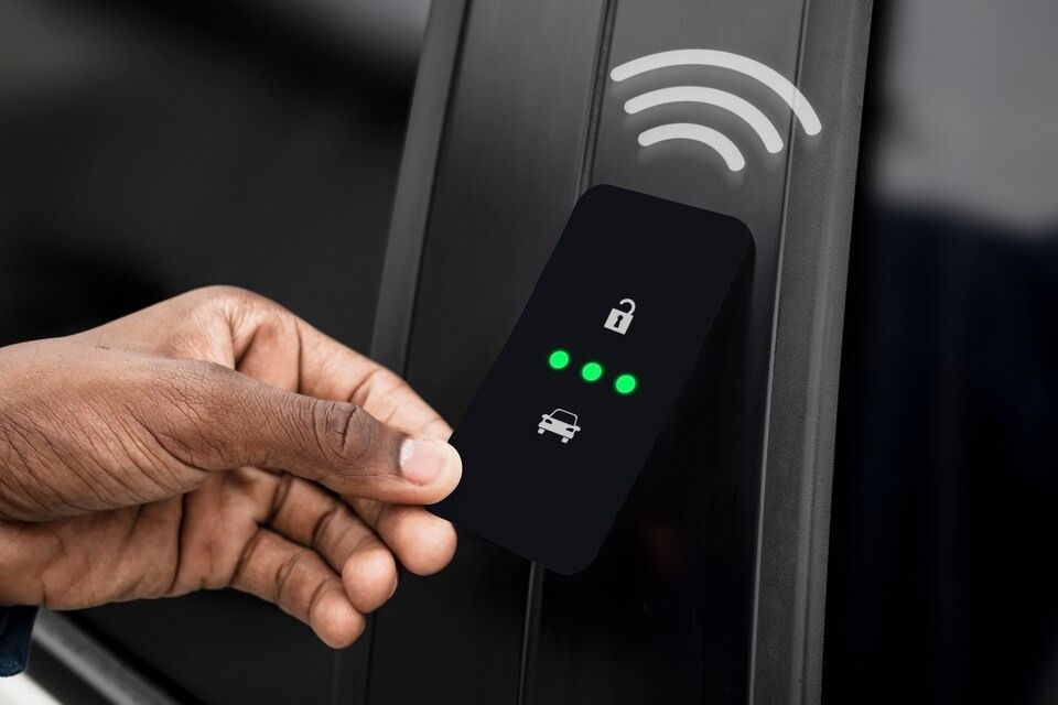 Person's hand holds black key card to unlock a car, with green lights indicating access. Wireless signal shown.