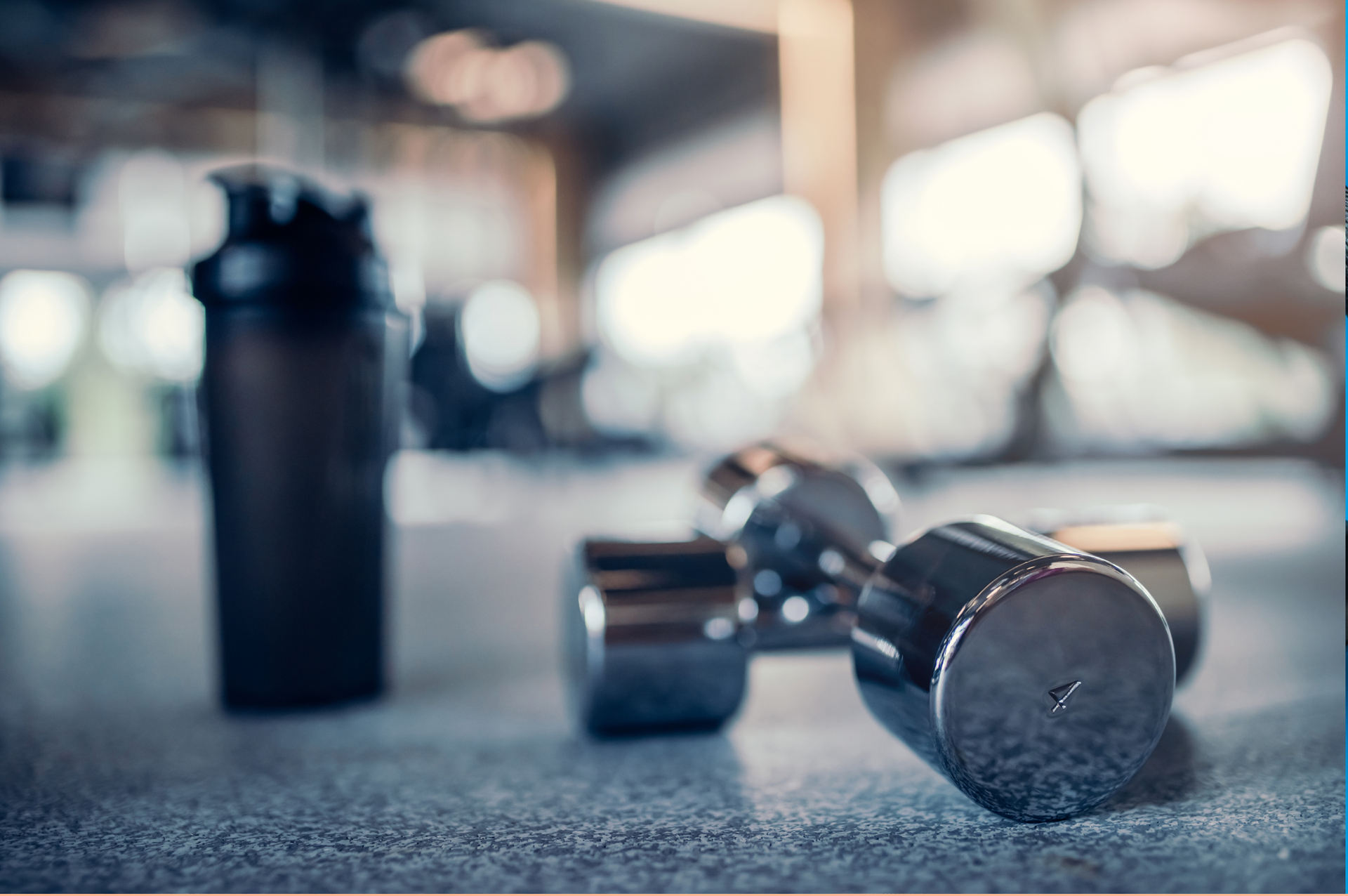 Manubri e una bottiglia d'acqua sul pavimento di una palestra, con attrezzi da allenamento sfocati sullo sfondo.