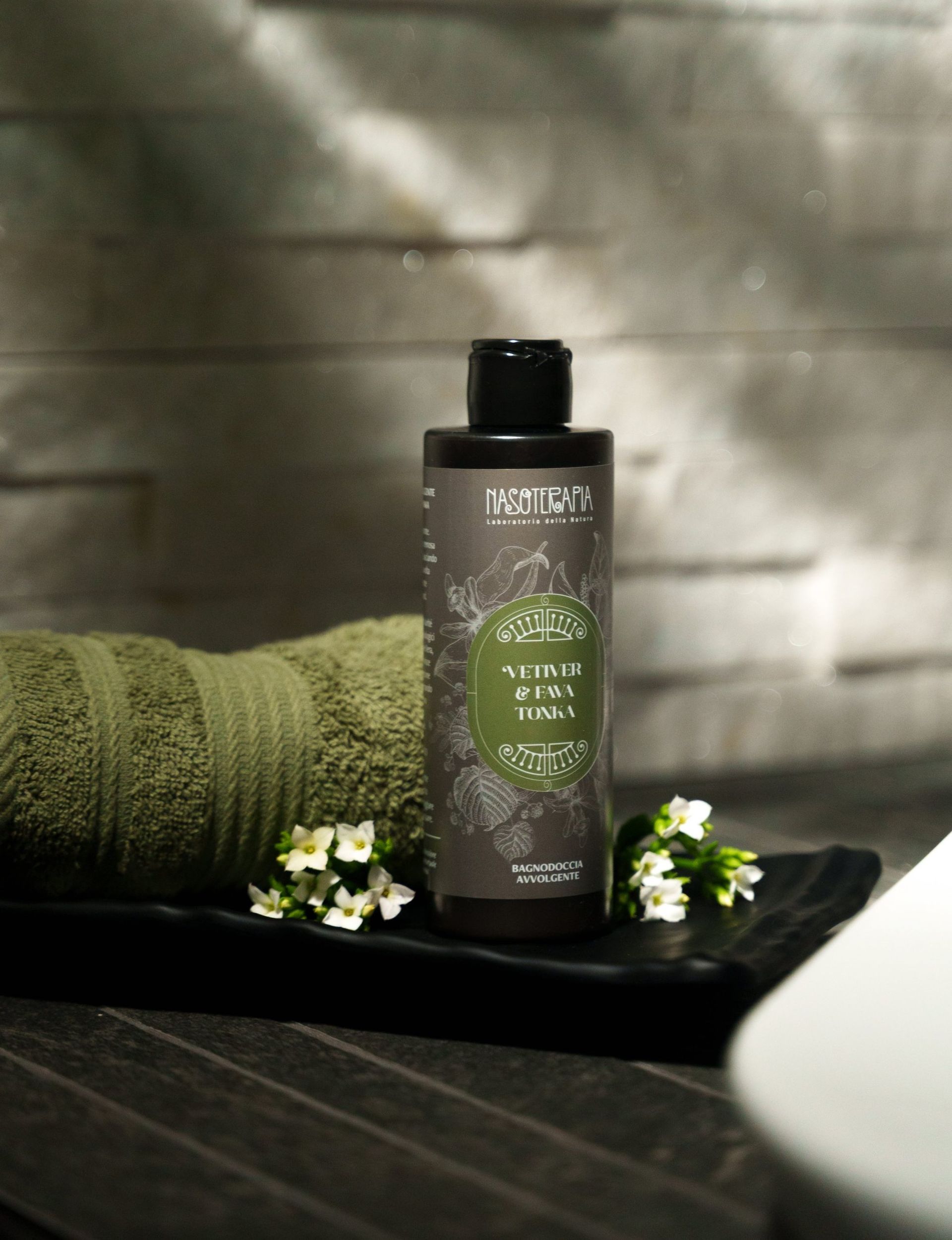 Flacone nero di shampoo su un vassoio con asciugamano verde e piccoli fiori bianchi.