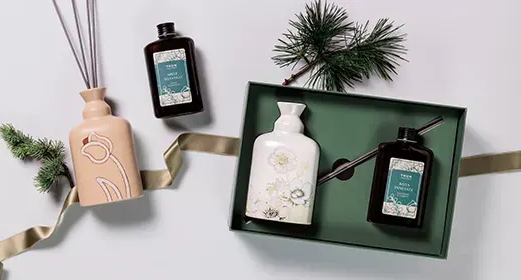 Set decorativo per il bagno con flaconi di lozione, diffusore a bastoncini, rametto di pino e nastro su sfondo chiaro.