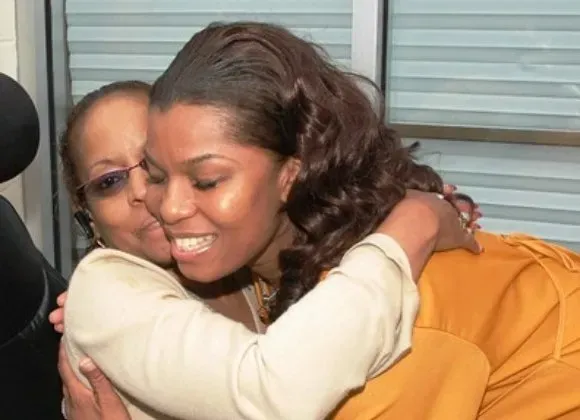 Two people hugging warmly indoors, smiling in front of a window blind.
