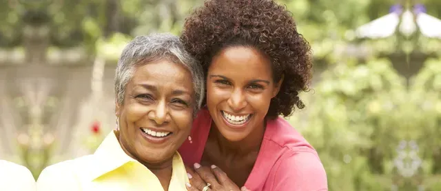 Two smiling people leaning together outdoors, with blurred greenery in the background