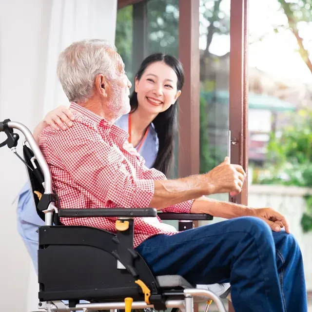 Smiling caregiver talking with a seated man in a wheelchair by a window
