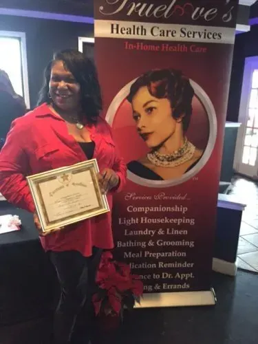 Woman in red shirt holding a framed certificate beside a health care services banner.