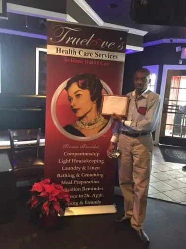 Man holding a framed certificate beside a True You’s Health Care Services banner and portrait display