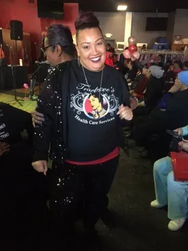 Smiling woman in a black sweatshirt at a crowded indoor event, with a red wall and balloon decorations behind her.