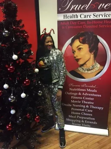 Woman posing beside a decorated Christmas tree and a TruLove Health Care Services banner.