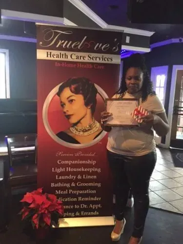 Woman holding a certificate beside a TrueBrew Health Care Services banner indoors