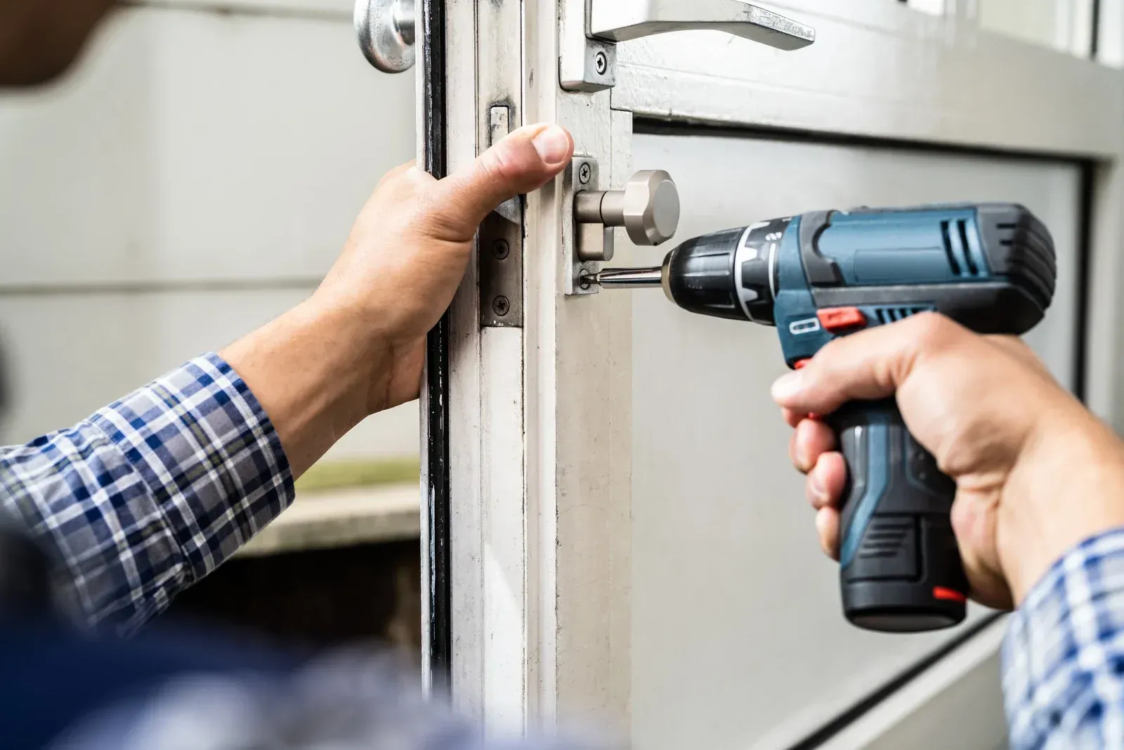 A person wearing a plaid shirt uses a cordless power drill to install a metal latch on a white door frame.