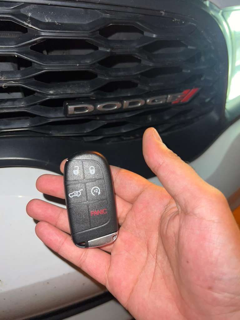 Hand holding a Dodge key fob in front of a Dodge grille and emblem