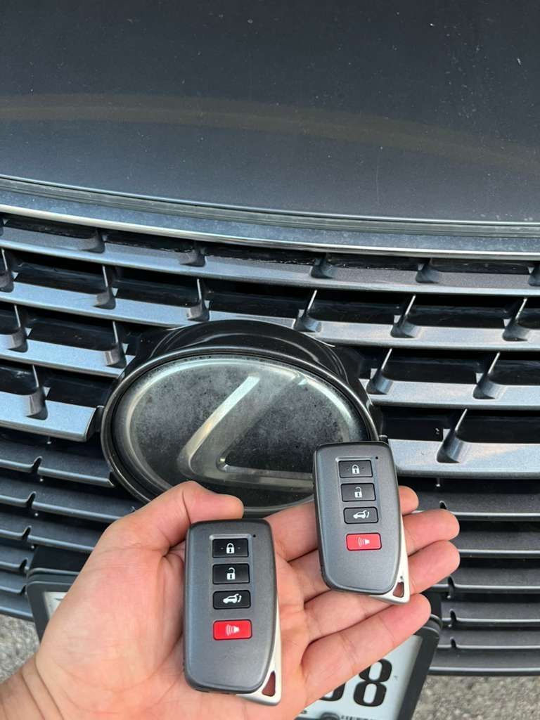 Hands holding two black car key fobs in front of a Lexus grille and emblem