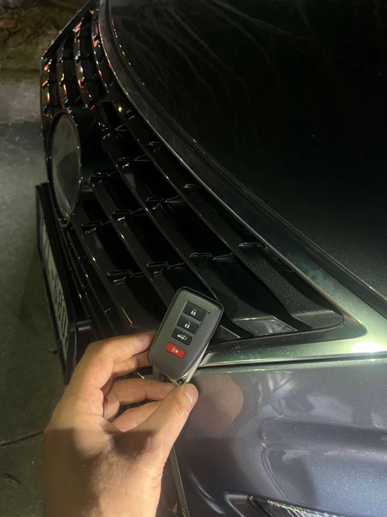 Hand holding a key fob near the front grille of a dark car at night