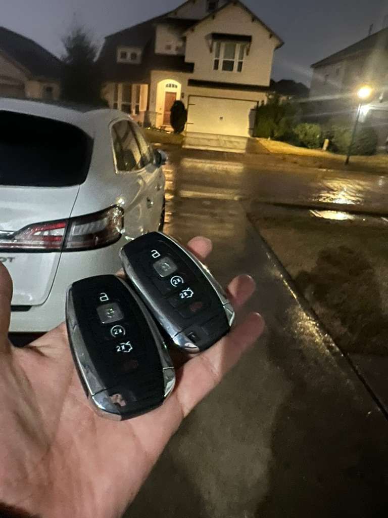 Hand holding two black car key fobs beside a white car on a wet driveway at night