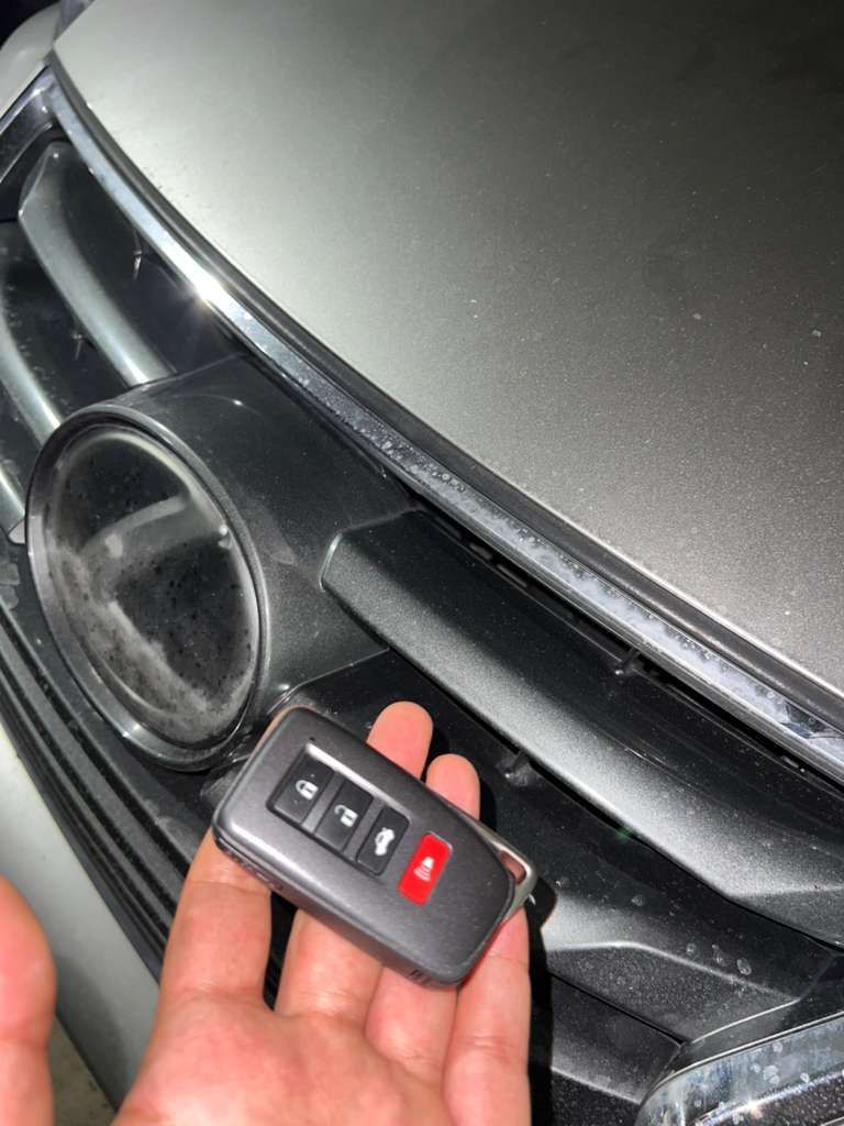 Hand holding a black car key fob near the front grille and headlight of a silver car